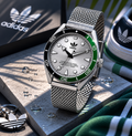 Fotografía de producto del reloj deportivo Adidas Edition Two sobre bloques de hormigón con gotas de agua. El reloj destaca por su brazalete de malla milanesa plateada, esfera gris cepillada y bisel giratorio bicolor en negro y verde. Al fondo se aprecian un llavero con el trébol de Adidas y parte de unas zapatillas a juego.