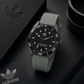 Reloj analógico deportivo Adidas Edition Two con correa de silicona gris claro y caja gris oscuro mate. El dial negro destaca con el logotipo del trébol y un bisel de buceo con números blancos. Se muestra sobre un fondo sofisticado junto a su estuche oficial de cuero.
