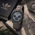 Fotografía de producto del reloj Adidas Originals AOFH22007 sobre una base de mármol. El reloj cuenta con caja redonda de 40 mm y correa de acero en color gris, dial gris con subesferas negras, manecilla azul eléctrico y bisel verde con escala taquimétrica. Al fondo se aprecia un cuaderno negro con el logotipo del trébol de Adidas.