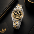 Fotografía de producto en primer plano del reloj Adidas Edition One AOFH23010. Destaca su caja de acero redonda con bisel interno dorado, esfera negra con un gran trébol de Adidas en oro amarillo y brazalete de eslabones bicolor plata y oro. Reposa sobre un estuche de cuero negro junto a una tarjeta con el logotipo dorado