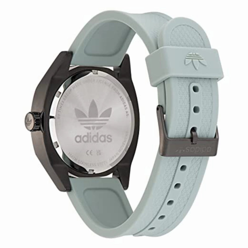 Reloj analógico deportivo Adidas Edition Two con correa de silicona gris claro y caja gris oscuro mate. El dial negro destaca con el logotipo del trébol y un bisel de buceo con números blancos. Se muestra sobre un fondo sofisticado junto a su estuche oficial de cuero. parte trasera